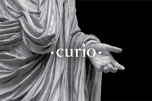 A While Curio logo overlaid onto a grey marble Statue of a robed figure with hand outreached against a black background.