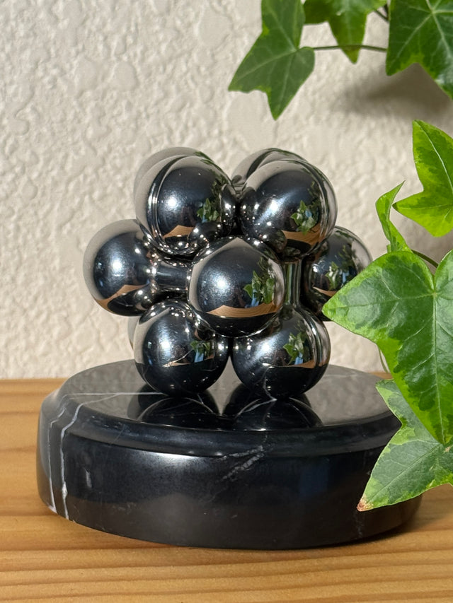 The silver Curio Atom Puzzle assembled on a dark marble Curio Plinth, lifestyle shelf shot with ivy plant