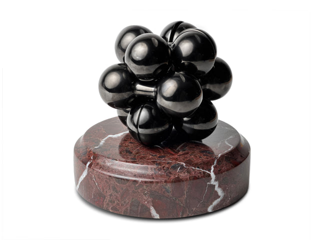 A black Curio Atom Puzzle resting on a Roso Lepanto Curio Plinth on a white background