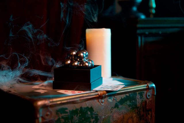 The assembled silver Curio Atom Puzzle resting on its gift box, styled with playing cards, a lit candle, and a vintage trunk, lifestyle shot