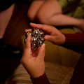 Two hands passing the assembled silver Curio Atom Puzzle between them, lifestyle shot with warm dim lighting