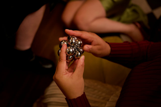 Two hands passing the assembled silver Curio Atom Puzzle between them, lifestyle shot with warm dim lighting