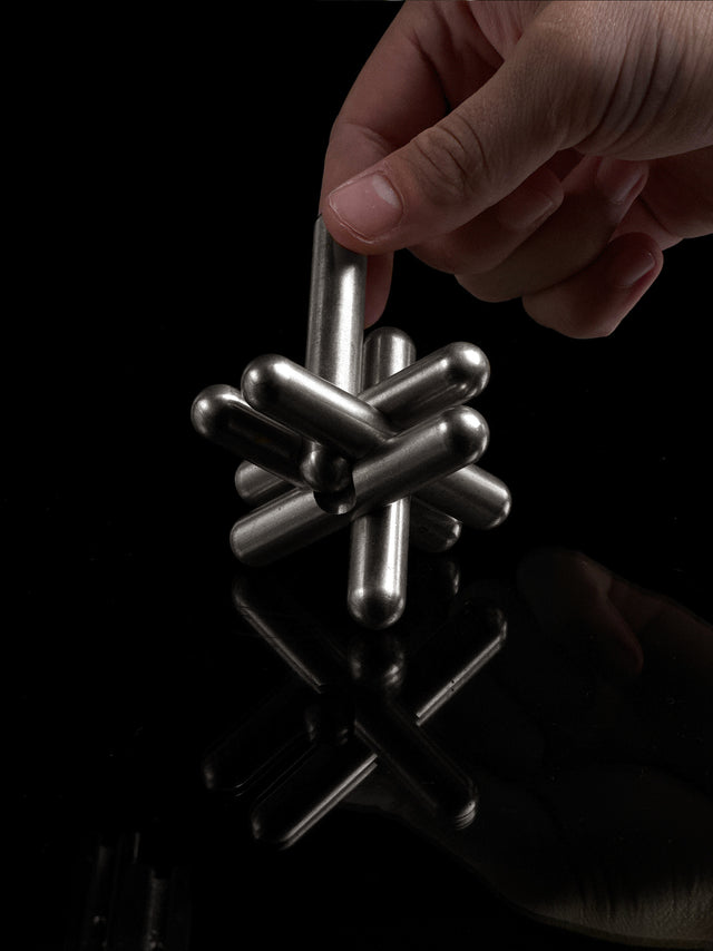 A hand pulling one bar from the assembled stainless steel Curio Jack Puzzle on a black reflective surface